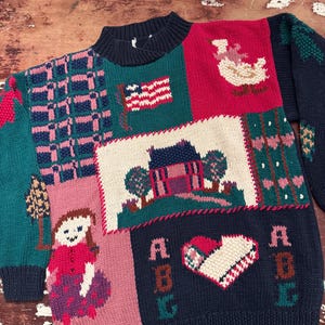 Könnte beinhalten: Ein farbenfroher Pullover im Patchwork-Stil mit einem marineblauen Rippkragen und -bündchen. Der Pullover zeigt verschiedene Strickmuster, darunter eine amerikanische Flagge, ein Haus, ein Mädchen, Herzen und die Buchstaben "ABC". Die Farbpalette umfasst Türkis, Rosa, Rot und Weiß.