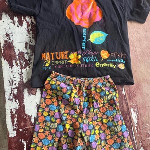 Può includere: Una t-shirt nera a maniche corte con una grande grafica di una rosa arancione e rosa, la parola ESPRIT e testo che include NATURE, seeds of hope, spirit ed Energy. Abbinata a pantaloncini con un motivo floreale multicolore.