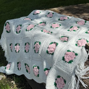May include: A white crochet blanket with a repeating pattern of pink roses and green leaves. The blanket has a fringe border and is draped over a wooden surface. The blanket is handmade and suitable for a bed or sofa.
