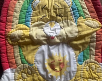 May include: A quilted fabric wall hanging featuring a yellow bear character with a sun emblem on its belly, positioned in front of a multi-colored rainbow arch. The bear has closed eyes, a white muzzle, and is set against a white base background.