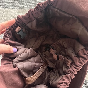 May include: A maroon-colored hat with a quilted interior. The hat features a drawstring closure and adjustable straps with black plastic buckles. The interior lining is a darker shade of brown, providing a contrast to the exterior.