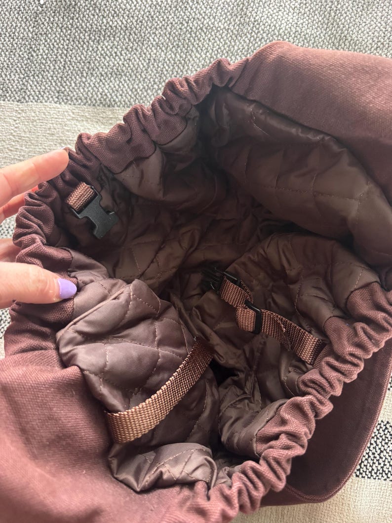 May include: A maroon-colored hat with a quilted interior. The hat features a drawstring closure and adjustable straps with black plastic buckles. The interior lining is a darker shade of brown, providing a contrast to the exterior.