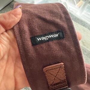 May include: Close-up of a dusty rose-colored dog accessory with a black label that reads "wagwear". The accessory has a brown strap and a black buckle. The fabric appears to be a textured canvas material.