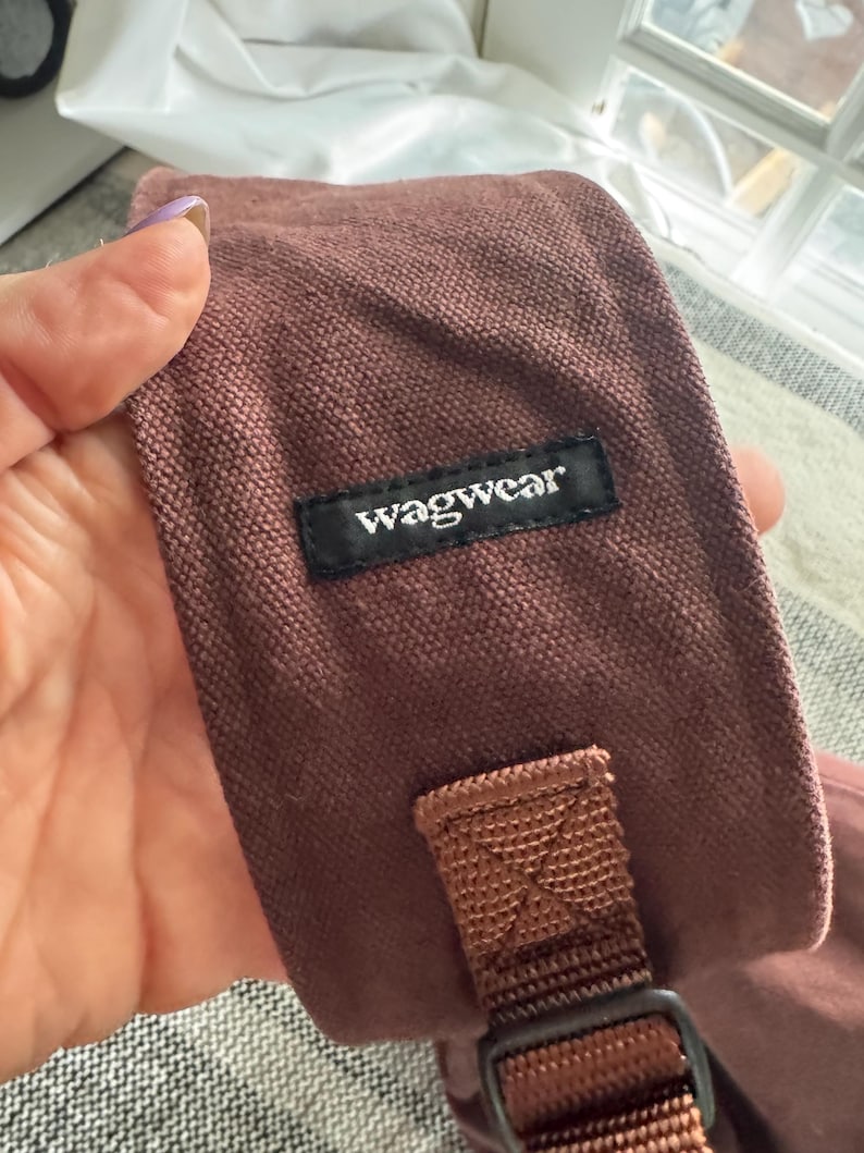 May include: Close-up of a dusty rose-colored dog accessory with a black label that reads "wagwear". The accessory has a brown strap and a black buckle. The fabric appears to be a textured canvas material.