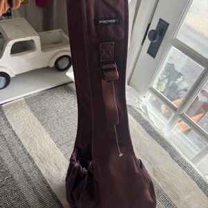 May include: A brown pet carrier sling with the brand name "wagwear" printed on the strap. The carrier has a drawstring closure and an adjustable strap with a buckle. The background includes a white toy truck and a window.