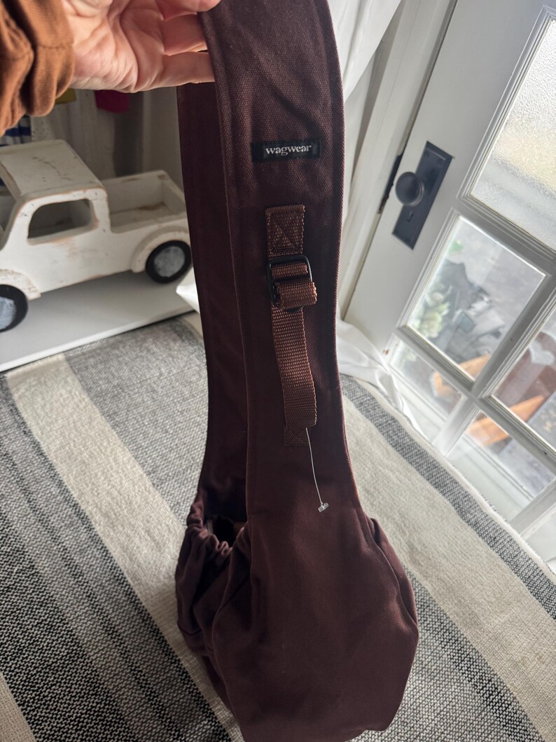 May include: A brown pet carrier sling with the brand name "wagwear" printed on the strap. The carrier has a drawstring closure and an adjustable strap with a buckle. The background includes a white toy truck and a window.