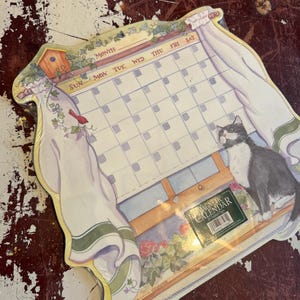 May include: A magnetic calendar featuring a grid for days of the week, illustrated with a black and white cat sitting by a window with white curtains, a birdhouse, and flowers. The text reads MAGNETIC CALENDAR 18 Undated Pages.