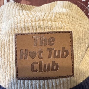 Könnte beinhalten: Cord-Baseballkappe in Creme und Marineblau. Auf der Vorderseite befindet sich ein braunes Lederetikett mit dem geprägten Schriftzug "The Hot Tub Club" und einem Herzsymbol. Die Kappe hat ein klassisches, strukturiertes Design.