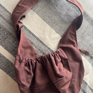 May include: A dusty rose-colored sling bag with a wide shoulder strap. The bag has a gathered top and a small pocket. A thin strap with a metal clasp is attached. The bag is made of a soft, possibly canvas-like material.