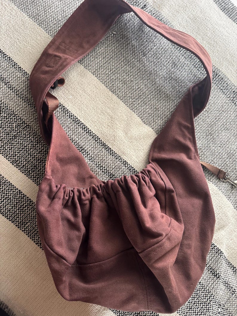 May include: A dusty rose-colored sling bag with a wide shoulder strap. The bag has a gathered top and a small pocket. A thin strap with a metal clasp is attached. The bag is made of a soft, possibly canvas-like material.