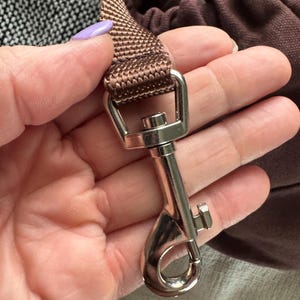 May include: A close-up of a brown nylon strap with a silver-colored metal clasp. The strap is looped and attached to a silver metal swivel hook. The background includes a brown fabric and a patterned surface.
