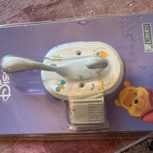Könnte beinhalten: Ein einzelner Disney-Haken in einer transparenten Kunststoffverpackung. Der Haken hat einen silbernen Griff und eine weiße Basis, die mit Winnie Puuh und Bienen verziert ist. Die Verpackung ist lila mit dem Disney-Logo und dem Wort "SINGLE HOOK".