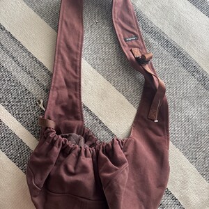 May include: A maroon-colored pet carrier bag with a long, wide shoulder strap. The bag has a drawstring closure at the top and a small pocket on the front. The bag is made of a soft, durable fabric.