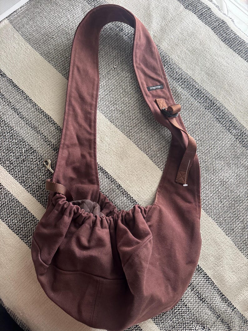 May include: A maroon-colored pet carrier bag with a long, wide shoulder strap. The bag has a drawstring closure at the top and a small pocket on the front. The bag is made of a soft, durable fabric.