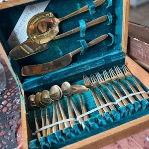 Könnte beinhalten: Ein Vintage-Besteckset in Goldtönen in einer Holzkiste mit türkisfarbenem Samtfutter. Das Set enthält Löffel, Gabeln, Messer und Servierutensilien. Die Box hat ein Etikett mit der Aufschrift "BANGKOK JEWELRY".