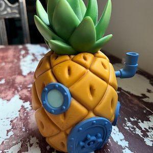 Puede incluir: Una figura con forma de casa de piña con un cuerpo amarillo texturizado, una parte superior de hojas verdes y detalles azules que incluyen una ventana circular, una puerta con manija en forma de rueda y un pequeño tubo azul lateral.