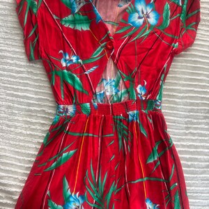 May include: Red romper with a tropical floral print featuring blue flowers and green leaves. The romper has short sleeves, a V-neckline, and a button closure. The waistline is elasticized.