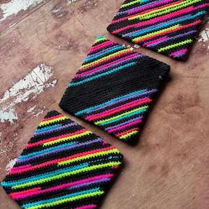 May include: Three square, hand-crocheted coasters. Each coaster features a black background with vibrant horizontal stripes in neon pink, yellow, green, blue, and purple. The coasters are arranged on a weathered wooden surface.