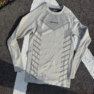May include: A light gray long-sleeve shirt featuring a crew neckline and a black geometric pattern across the chest and sides. The word YOUNGLA is printed in black on the upper chest area. The garment has a fitted silhouette with long sleeves.