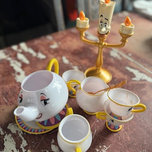 Könnte beinhalten: Ein Teeservice aus Kunststoff mit einer weißen Teekanne mit Gesicht, einer goldfarbenen Kandelaber-Figur mit drei orangefarbenen Flammen, einem weißen Milchkännchen und mehreren kleinen weißen Teetassen mit gelben Henkeln.