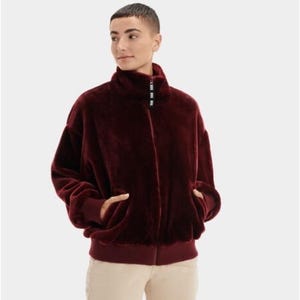 May include: A deep burgundy faux fur jacket featuring a high collar with a central zipper closure. The zipper pull is black with white text. The jacket has long sleeves with ribbed cuffs and a ribbed hem, providing a structured fit.