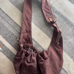May include: A maroon-colored pet carrier sling bag with a wide shoulder strap. The bag has a drawstring closure and a small pocket on the front. The strap has a buckle for adjustment. The bag is made of a soft, durable fabric.
