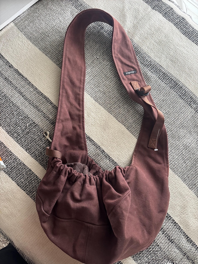 May include: A maroon-colored pet carrier sling bag with a wide shoulder strap. The bag has a drawstring closure and a small pocket on the front. The strap has a buckle for adjustment. The bag is made of a soft, durable fabric.