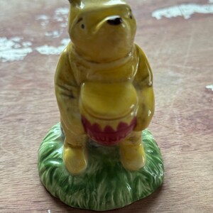 Classic" or "A.A. Milne" book-style depiction of Winnie the Pooh standing on a patch of green grass while clutching a pinkish-red pot