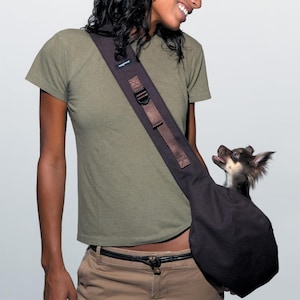 May include: A dark brown dog carrier sling with a small dog inside. The sling has a wide shoulder strap with a buckle and the word "wagwear" printed on it. The dog is looking up with its mouth open.