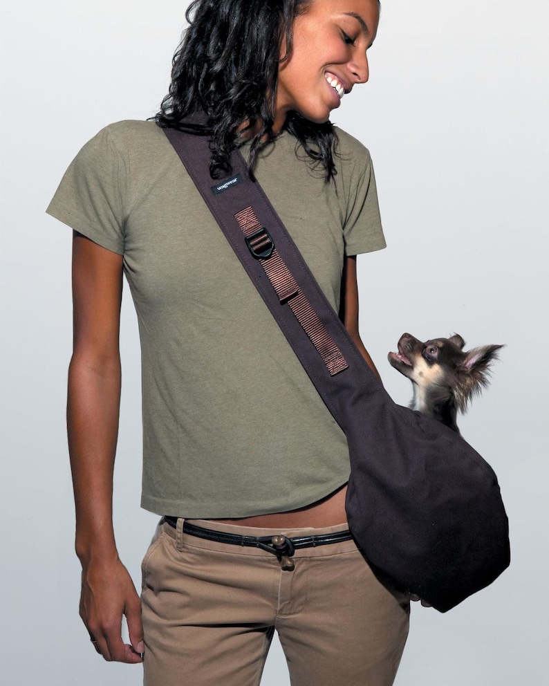 May include: A dark brown dog carrier sling with a small dog inside. The sling has a wide shoulder strap with a buckle and the word "wagwear" printed on it. The dog is looking up with its mouth open.