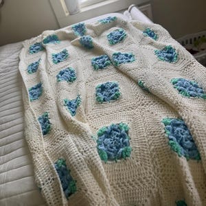 May include: Cream-colored crochet blanket with square floral patterns in shades of blue and green. The blanket is draped over a bed, showcasing its handmade texture and design. A cozy and decorative home textile.
