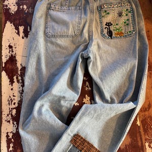 Könnte beinhalten: Hellblaue Jeans mit elastischem Bund und zwei Gesäßtaschen. Eine Gesäßtasche hat ein gesticktes Design einer schwarzen Katze, Pflanzen und farbigen Perlen. Die Vorderseite der Jeans hat ein Patchwork-Design mit einem braunen Karopatch.