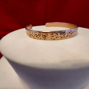 May include: A copper-toned metal cuff bracelet featuring an open-ended design and a textured surface with irregular, dark, etched markings across the exterior band. The bracelet has a polished finish and a consistent width throughout its circular form.