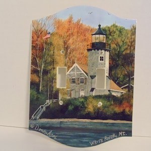May include: A light switch cover featuring a painting of a lighthouse with a white flag on a pole, surrounded by trees in autumn colors. The text "White River, ME." is at the bottom of the image.