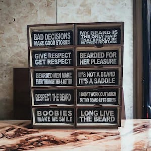 Pode incluir: Uma caixa de armazenamento de madeira com várias gavetas, cada uma exibindo um sinal retangular preto com texto branco. Os sinais apresentam citações humorísticas sobre barbas, como "Bad Decisions Make Good Stories" e "My Beard Is The Only Hair That Should Be Between Your Legs".