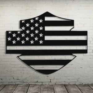 Op de afbeelding: Zwarte metalen wandkunst in de vorm van een schild, met het ontwerp van de Amerikaanse vlag. De sterren en strepen zijn uitgesneden, waardoor een negatief ruimte-effect ontstaat. De kunst is gemonteerd op een witte bakstenen muur met een houten vloer.