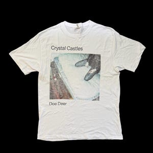 May include: White t-shirt with the text "Crystal Castles" above a rectangular image of a person lying on a mattress. The text "Doe Deer" is below the image. The shirt is made of a soft material.