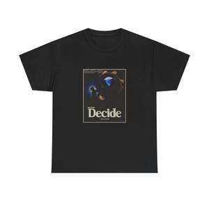 以下が含まれることがあります： 映画「Decide」のグラフィックがプリントされた黒いTシャツ。デザインは濃い青とオレンジの要素を含み、白で「Decide」の文字が書かれています。Tシャツはクルーネックで、クラシックなスタイルです。