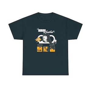 Puede incluir: Camiseta de color verde azulado oscuro con un diseño gráfico con el texto "Birds of a Feather". El diseño incluye un retrato de una mujer, estrellas y otras ilustraciones en blanco y amarillo. Una camiseta unisex informal.