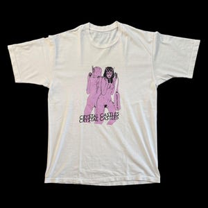 May include: Off-white t-shirt featuring a pink graphic of two figures with the text "CRYSTAL CASTLES" below. The shirt has short sleeves and a crew neck. The graphic includes the text "AFFECTION EP".