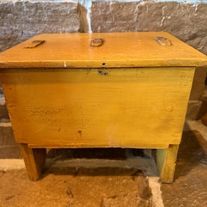 May include: A rectangular wooden box with a mustard yellow finish, featuring a flat lid with three small metal hinges along the top edge. The box is supported by two sturdy wooden legs and displays a visible wood grain texture throughout its surface.