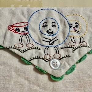 May include: Embroidered white fabric with a green scalloped edge. The fabric features three whimsical, anthropomorphic embroidered figures: a red teacup, a blue circle, and a yellow spiral, each with legs and smiling faces. A white button is sewn on the fabric.