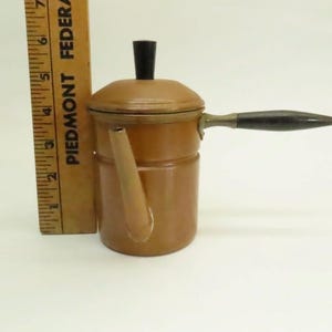 May include: A small, tan-colored coffee pot with a black handle and lid knob. The pot has a spout and is next to a wooden ruler. The ruler is marked with the text "PIEDMONT FEDERAL".