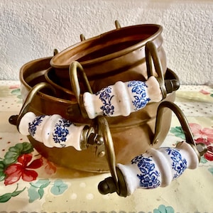 May include: A set of three copper pots nested together. Each pot has a brass handle and white ceramic handles with blue floral designs. The pots are arranged on a floral patterned surface.
