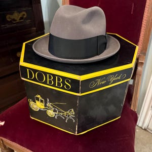 May include: A grey fedora hat with a black ribbon band resting on a hexagonal black and yellow hat box. The box features the text DOBBS New York and a graphic illustration of a horse-drawn carriage in yellow and white on the side panels.