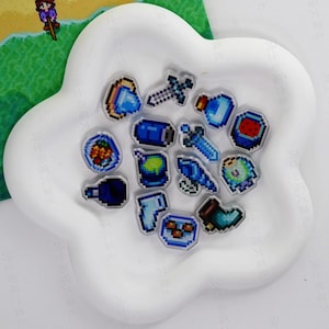 May include: A selection of pixel art acrylic charms, featuring game-related icons such as swords, potions, and food. The charms are in blue, green, and red hues, displayed on a white, flower-shaped dish.