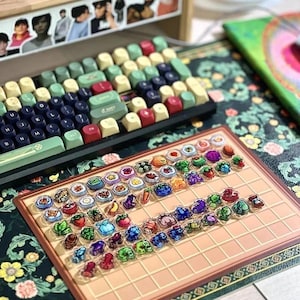 May include: A mechanical keyboard with keycaps in green, red, and beige sits above a tray of decorative food-themed keycaps. The keyboard rests on a floral patterned surface.