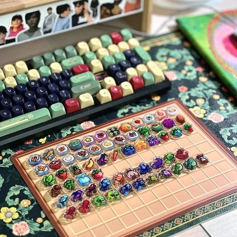 May include: A mechanical keyboard with keycaps in green, red, and beige sits above a tray of decorative food-themed keycaps. The keyboard rests on a floral patterned surface.