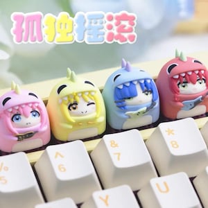May include: A set of four colorful anime-style keycaps, each in a different color: pink, yellow, blue, and red. The keycaps are shaped like cartoon characters with dinosaur-like hoods. The keycaps are on a keyboard with white keys.