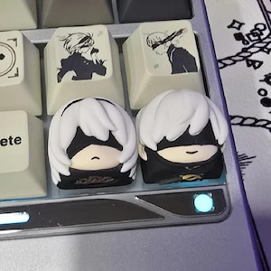 May include: Two custom artisan mechanical keyboard keycaps featuring sculpted 3D character heads with white hair and black blindfolds. Behind them are two flat keycaps with black and white printed illustrations of the same characters.